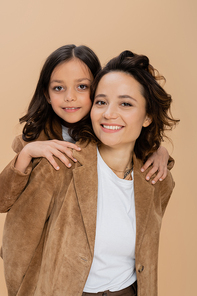 brunette girl hugging shoulders of happy mom in brown suede jacket isolated on beige