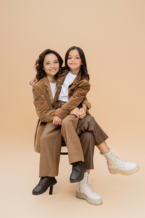 happy mother and daughter in trendy autumn outfit and boots sitting on chair on beige