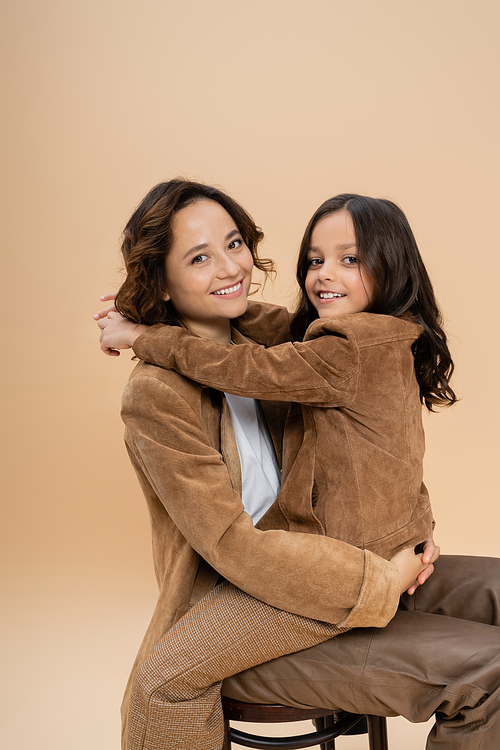 happy mother and daughter in trendy autumn clothes embracing and looking at camera isolated on beige