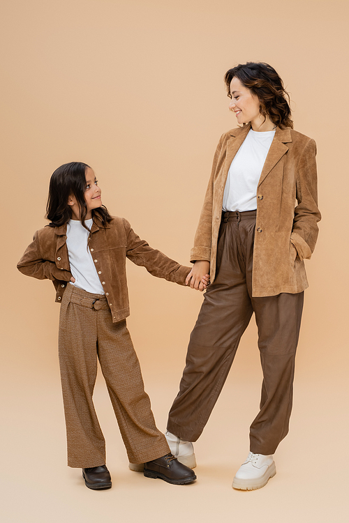 full length of woman and girl in trendy autumn outfit holding hands and smiling at each other on beige background