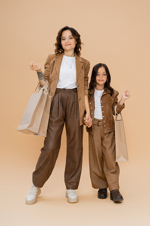 full length of woman and girl in stylish autumn clothes standing with shopping bags on beige background