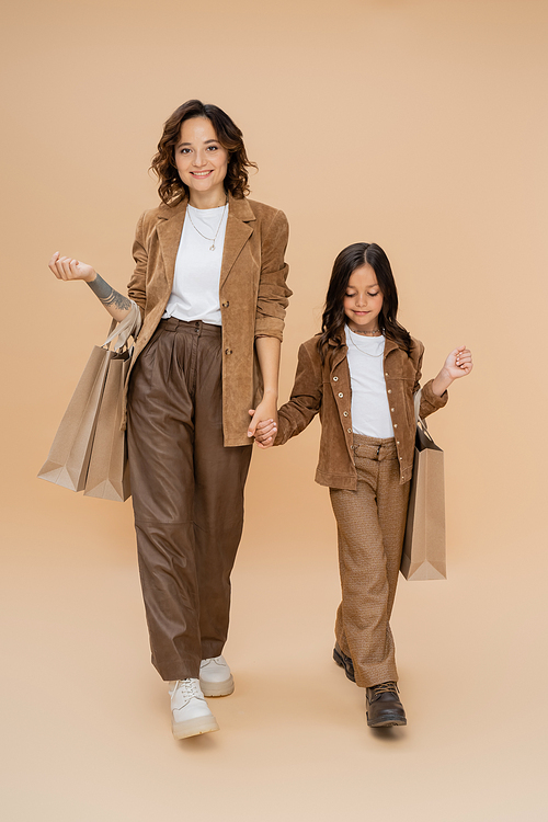 mother and child in trendy autumn clothes holding hands while walking with shopping bags on beige