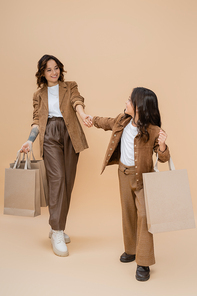 trendy girl with shopping bags holding hands with cheerful and stylish mom on beige background