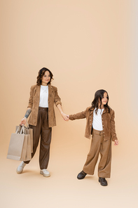 full length of girl in trendy clothes holding hands with happy mom with shopping bags on beige background