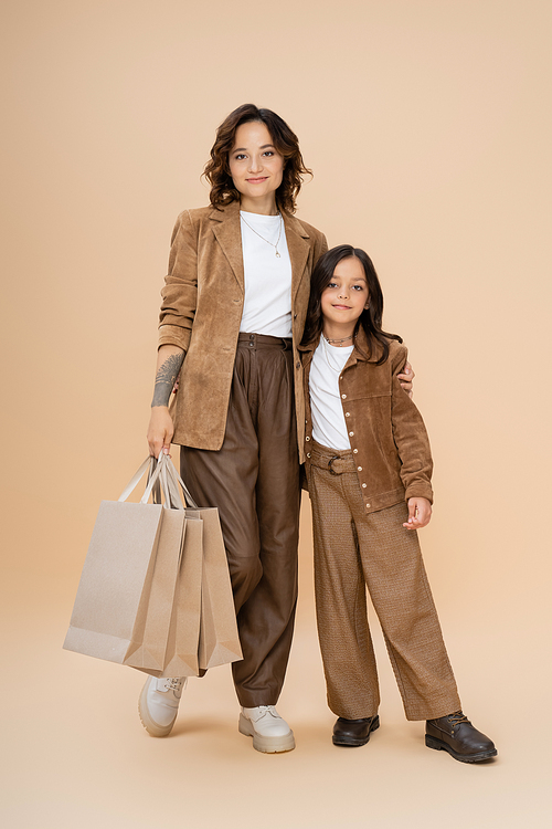 full length of woman with shopping bags hugging daughter while posing in trendy autumn outfit on beige