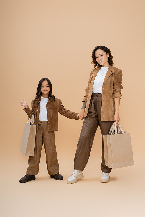 happy mom and daughter in trendy autumn outfit holding hands and shopping bags on beige