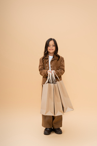 smiling girl in trendy autumn clothes holding shopping bags and looking at camera on beige background