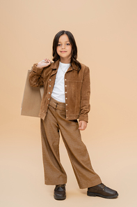 brunette girl in suede jacket and brown pants posing with shopping bags on beige background