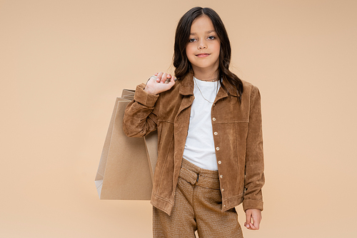 brunette girl with shopping bags posing in suede jacket and looking at camera isolated on beige