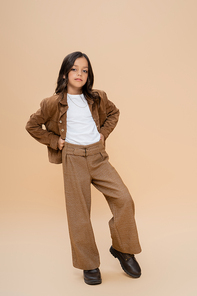 full length of trendy girl in brown pants and suede jacket standing with hands on hips on beige background