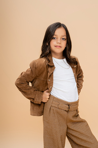 brunette girl in brown suede jacket posing with hands on hips while looking at camera isolated on beige