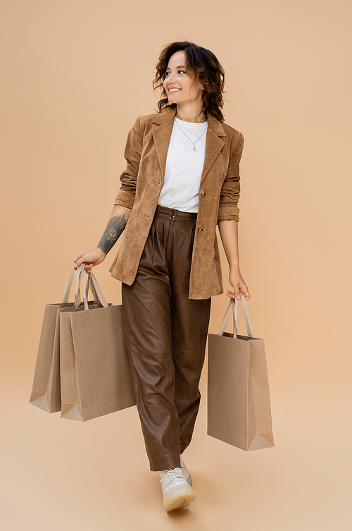 joyful woman in suede jacket walking with shopping bags and looking away on beige background