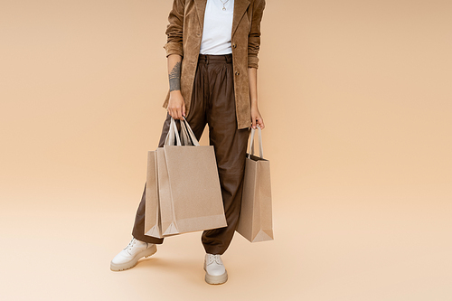 partial view of tattooed woman in brown pants holding shopping bags on beige background