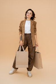 full length of woman in trendy autumn outfit posing with shopping bags on beige background