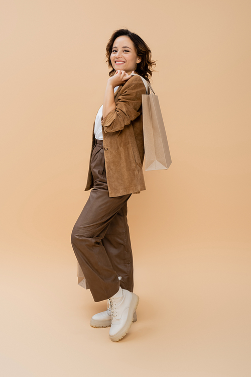 full length of smiling woman in brown pants and suede jacket holding shopping bag on beige