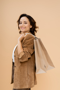 brunette woman in brown suede jacket holding shopping bags while smiling at camera isolated on beige