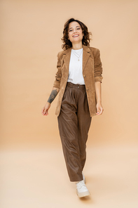 full length of tattooed woman in suede jacket and brown pants walking on beige