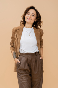 young woman in suede jacket standing with hands in pockets of brown pants isolated on beige