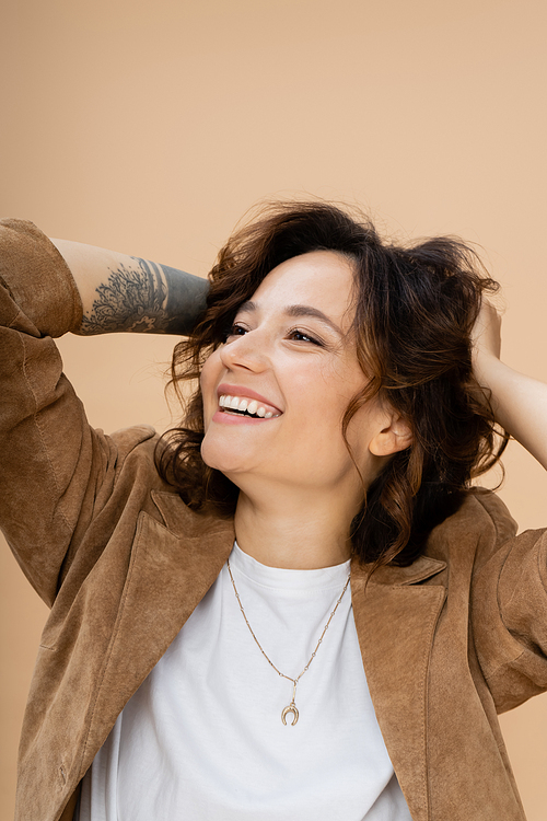 happy tattooed woman in brown suede jacket touching wavy hair and looking away isolated on beige