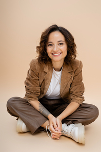 cheerful tattooed woman in suede jacket and trendy pants and boots sitting with crossed legs on beige background
