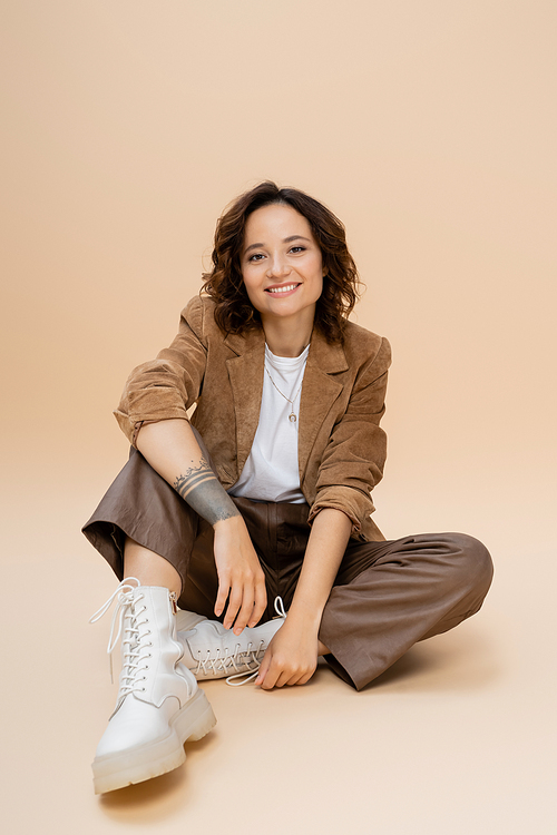 brunette woman in boots and trendy autumn clothes sitting on beige background