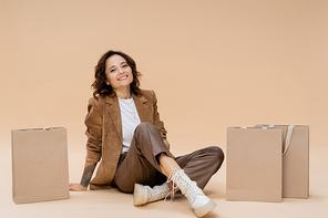 smiling woman in suede jacket and brown pants sitting near shopping bags on beige background