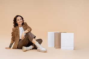 happy woman in stylish autumn clothes and boots sitting near shopping bags on beige background