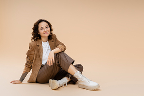 smiling woman in suede jacket and stylish pants and boots sitting on beige background