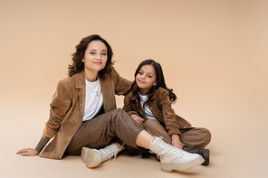 happy woman in brown jacket and trendy boots embracing daughter while sitting on beige background
