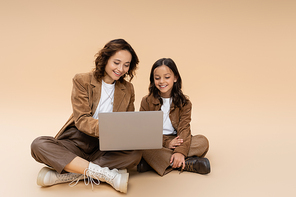 smiling girl with mom in stylish autumn clothes sitting with crossed legs near laptop on beige background