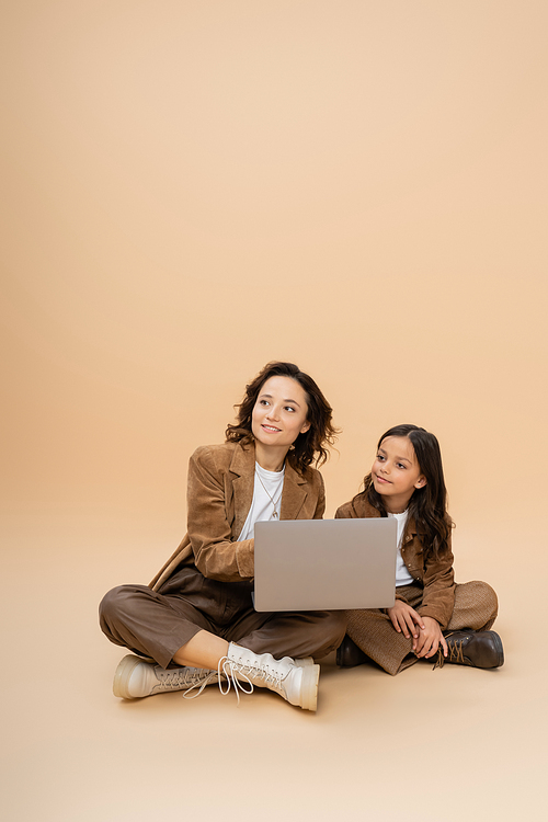 smiling and stylish woman looking away near laptop and daughter on beige background