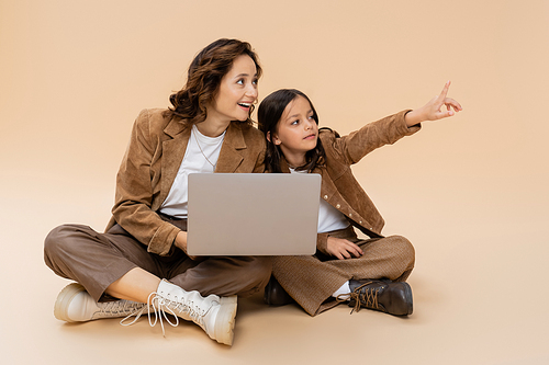 girl in trendy autumn clothes pointing with finger near laptop and smiling mom on beige background