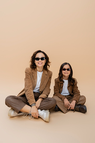 cheerful girl and mother in stylish autumn outfit and sunglasses sitting with crossed legs on beige background