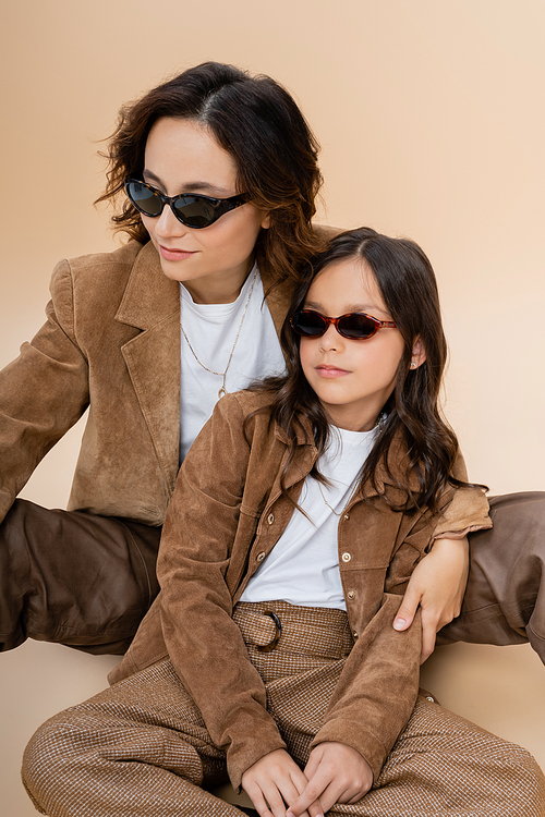 fashionable mother and daughter in sunglasses and brown suede jackets sitting on beige background