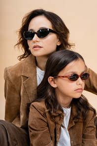 trendy mother and daughter in sunglasses and brown suede jackets isolated on beige