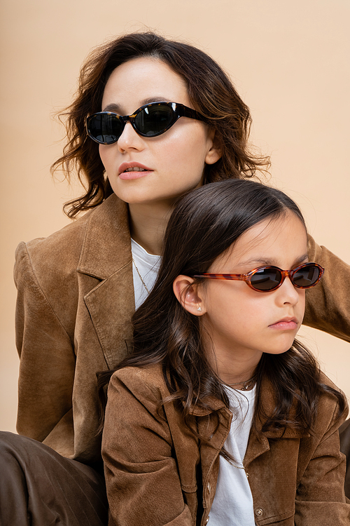 trendy mother and daughter in sunglasses and brown suede jackets isolated on beige