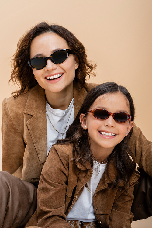 happy and stylish mother and daughter in suede jackets and sunglasses isolated on beige