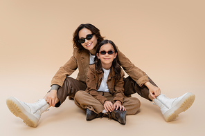 fashionable mom and daughter in sunglasses posing in suede jackets and boots on beige background