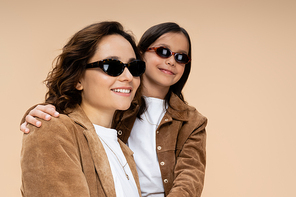 mother and child in suede jackets and stylish sunglasses smiling isolated on beige