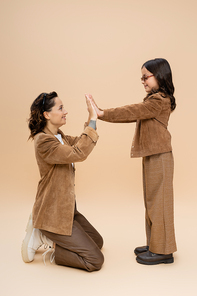 side view of mother and daughter in stylish autumn outfit playing patty cake game on beige background