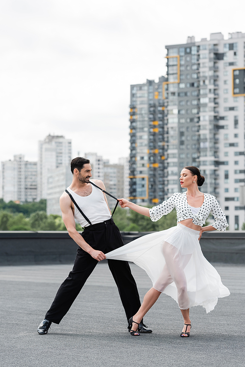 Stylish woman in heels dancing with smiling partner on roof of building outdoors