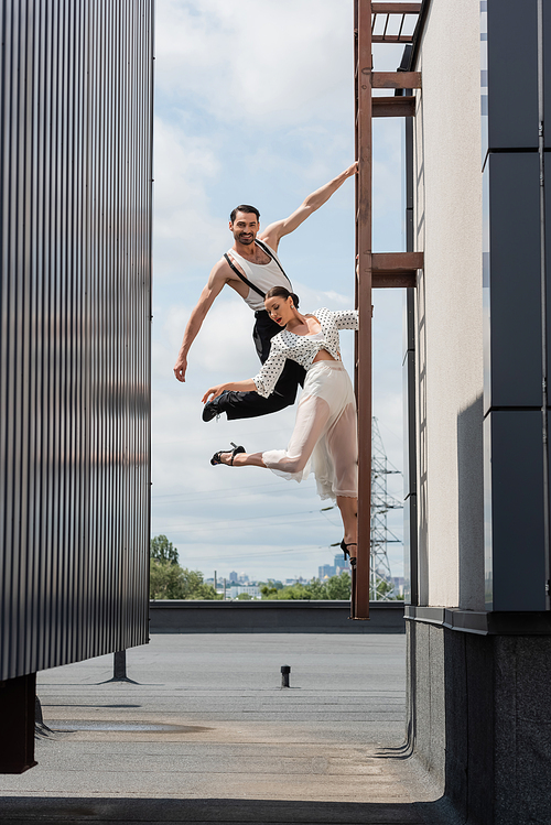 Smiling dancer looking at camera while posing near partner in skirt and heels on ladder on roof