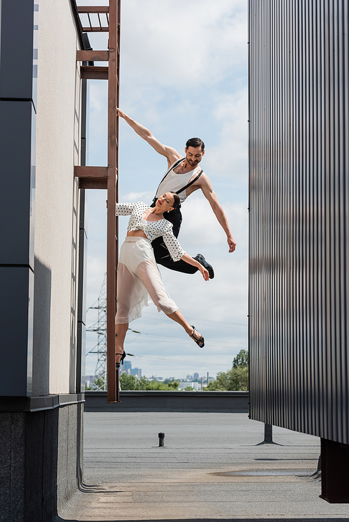 Smiling professional dancer looking at partner in heels posing on ladder on rooftop of building outdoors