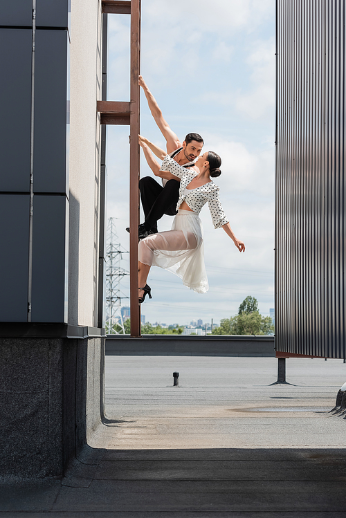 Professional ballroom dancers looking at each other while posing on ladder on rooftop of building outdoors