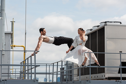 Stylish professional dancer looking at smiling partner on railing on building roof