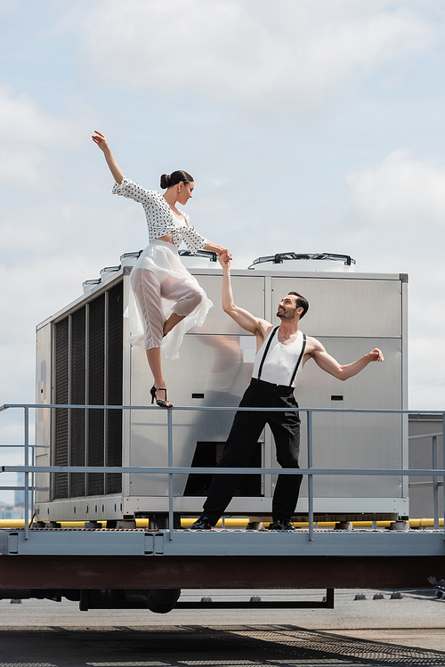 Smiling dancer in heels and skirt holding hand of partner on roof of building outdoors