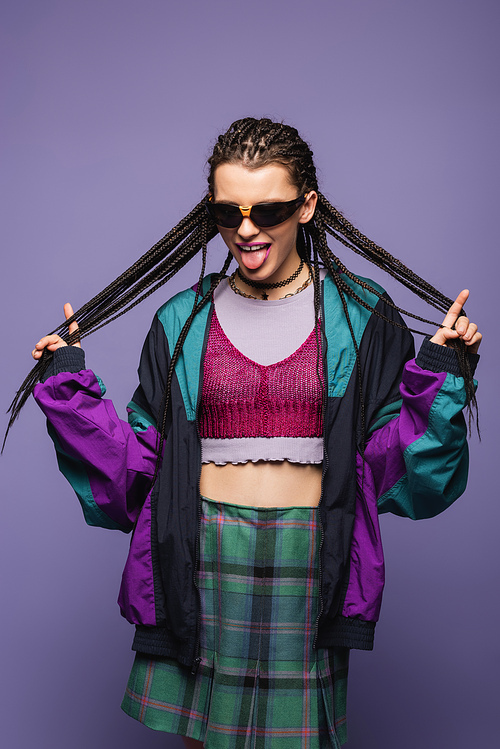 Stylish woman in vintage clothes touching braids and sticking out tongue isolated on purple
