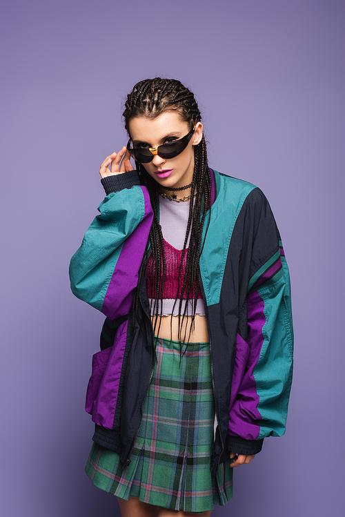 Young woman in retro sports jacket holding sunglasses on purple background