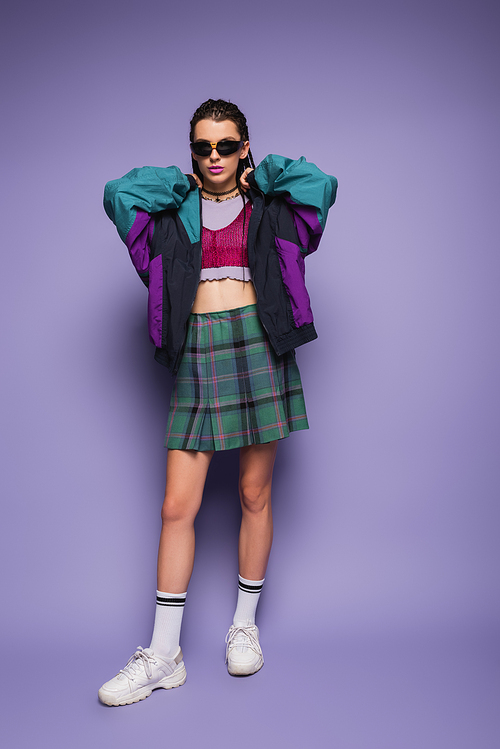 Trendy young model in plaid skirt and sports jacket standing on purple background