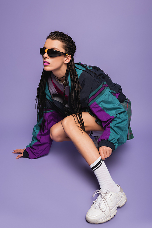 young woman in sunglasses and vintage jacket adjusting white sock on purple background